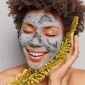 woman gets facial mask made natural herbal ingredients smiles toothily keeps eyes closed stands with bare shoulders enjoys beauty product poses indoor dermatology skin care 85x85